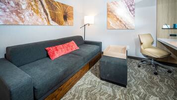 Una habitación de hotel moderna con un sofá gris, una almohada roja, una mesita de madera, una silla color beige y un escritorio con una lámpara.
