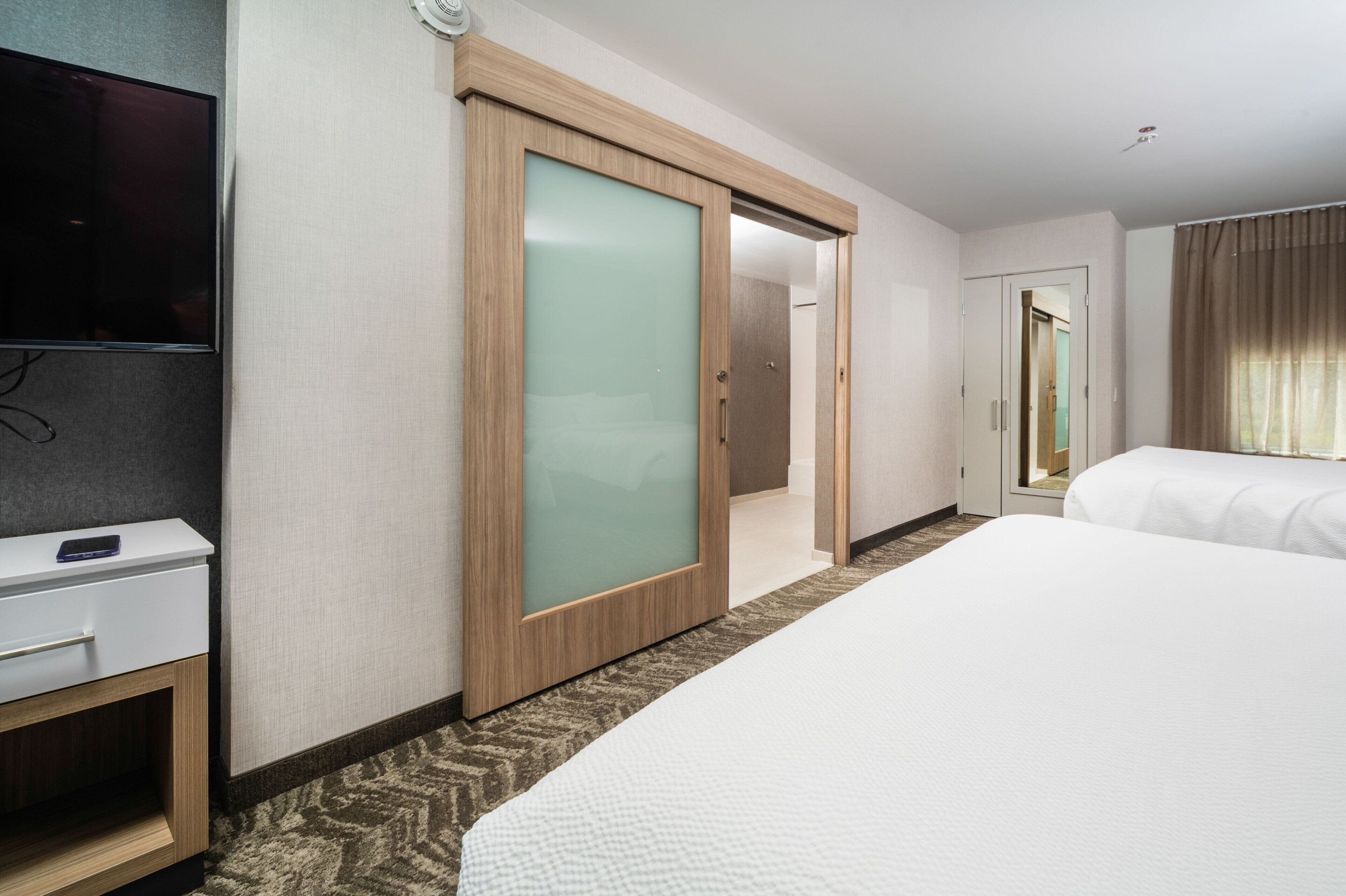 Quarto de hotel com uma cama, uma escrivaninha com telefone, uma porta de correr e uma televisão.
