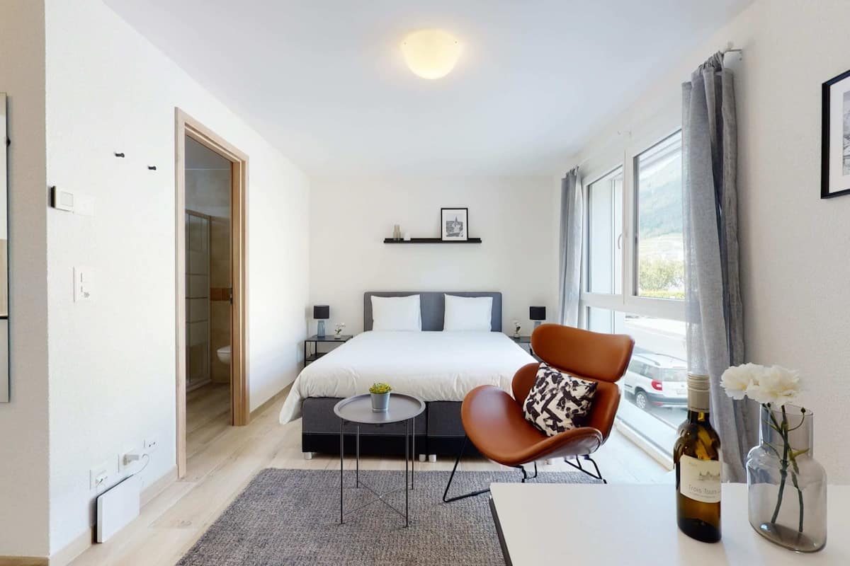 A modern hotel room with a large bed, a seating area with a chair and a small table, a bottle of wine, and a vase with flowers.