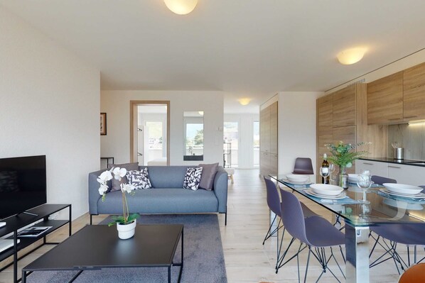 A modern living room with a sofa, a coffee table with a plant, a TV, and a dining area with a glass table and chairs.
