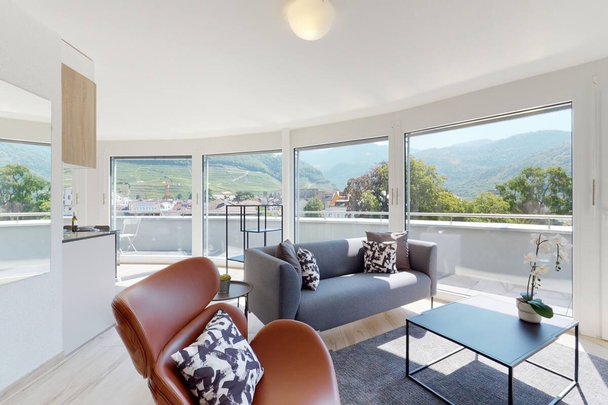 A modern living room with a grey sofa, brown leather armchair, and a small table. Large windows offer a view of a green landscape and distant buildings.