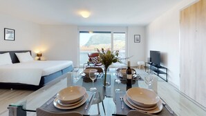 A modern hotel room with a bed, a dining table set for two, a TV, and a view of a mountainous landscape. - Nonanteneuf Aigle (Aigle)