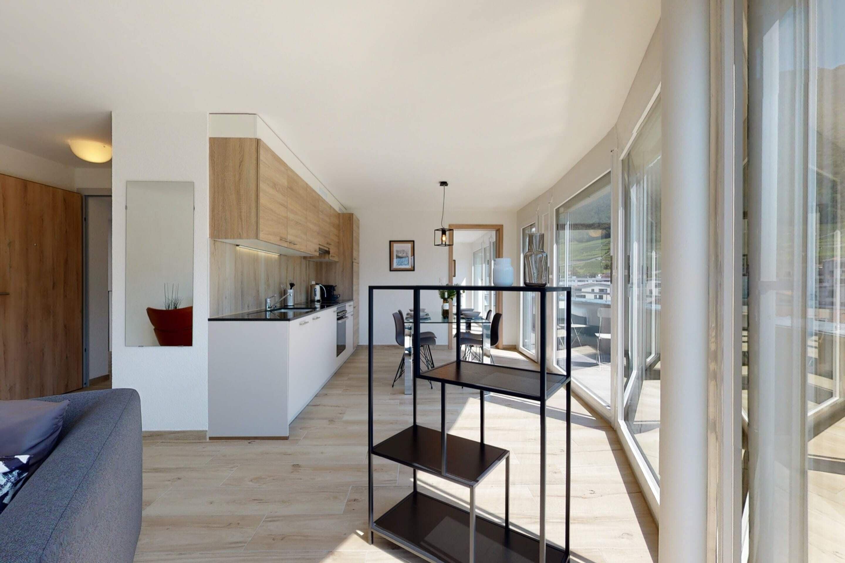 A modern kitchen with wooden cabinets, a dining area with a glass table, and a living room with a sofa.