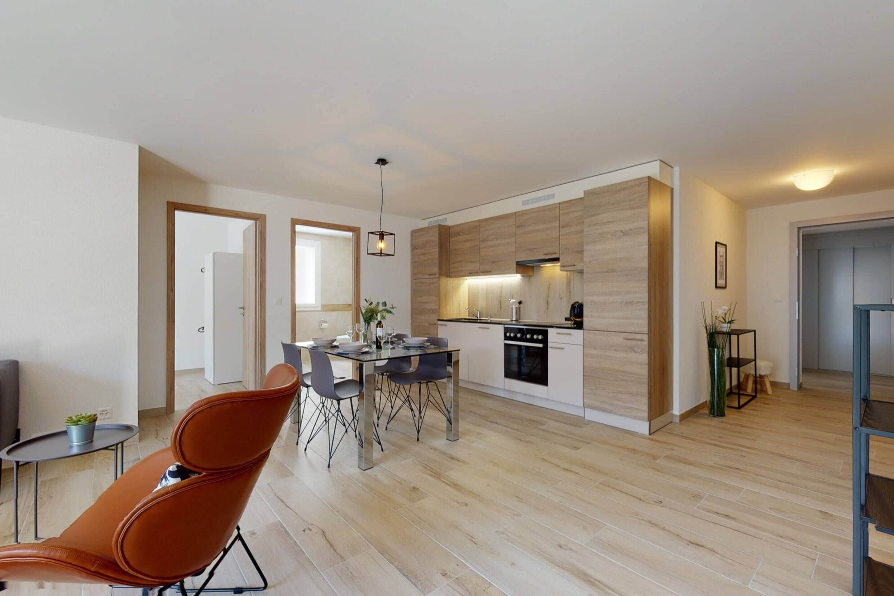 A modern kitchen with wooden cabinets, a dining area with a table and chairs, and a lounge chair.