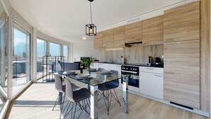 A modern kitchen with a glass dining table, wooden cabinets, and a view of the city. - Nonanteneuf Aigle (Aigle)