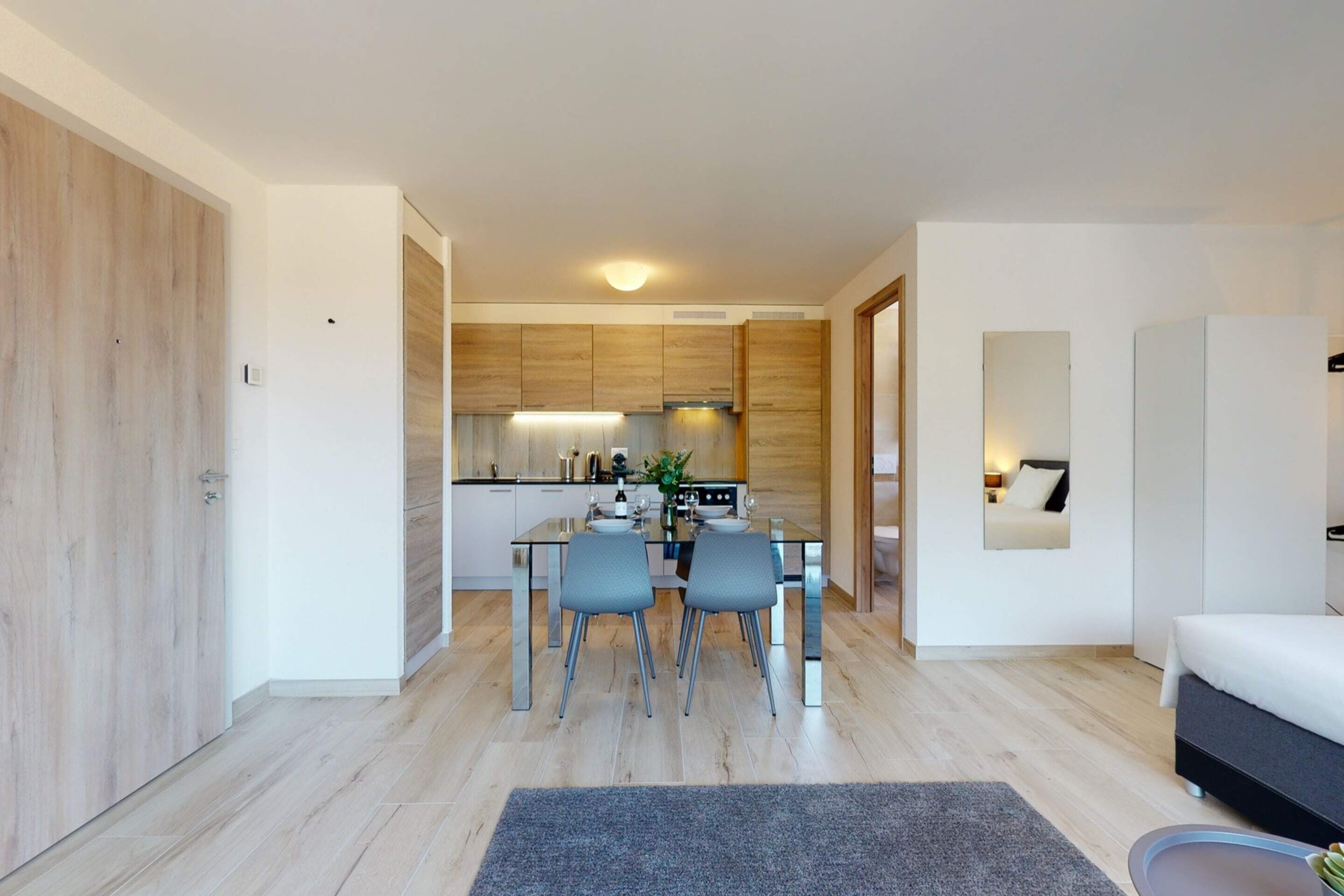Eine moderne Küche mit Holzschränken, ein Essbereich mit zwei Stühlen und ein Wohnzimmer mit Sofa und Couchtisch.