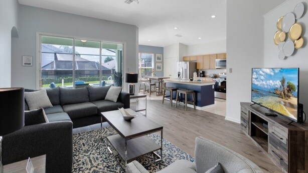 A modern living room with a sofa, coffee table, and TV. It includes a kitchen area with a bar and large windows. - Immaculate 4 Bedrooms 3 Bathroom In Windsor Palms Villa by RedAwning (Kissimmee)