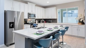 Una cocina moderna con armarios blancos, electrodomésticos de acero inoxidable, una isla central con taburetes y un amplio ventanal que proporciona luz natural.