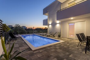 Un espace piscine extérieur moderne, doté d’une terrasse carrelée, de chaises longues et offrant une vue sur les toits de la ville au crépuscule.