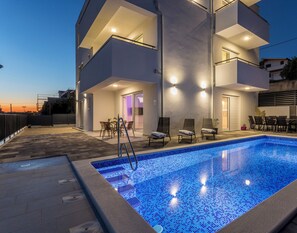A modern two-story building with a swimming pool, outdoor seating, and a well-lit entrance.