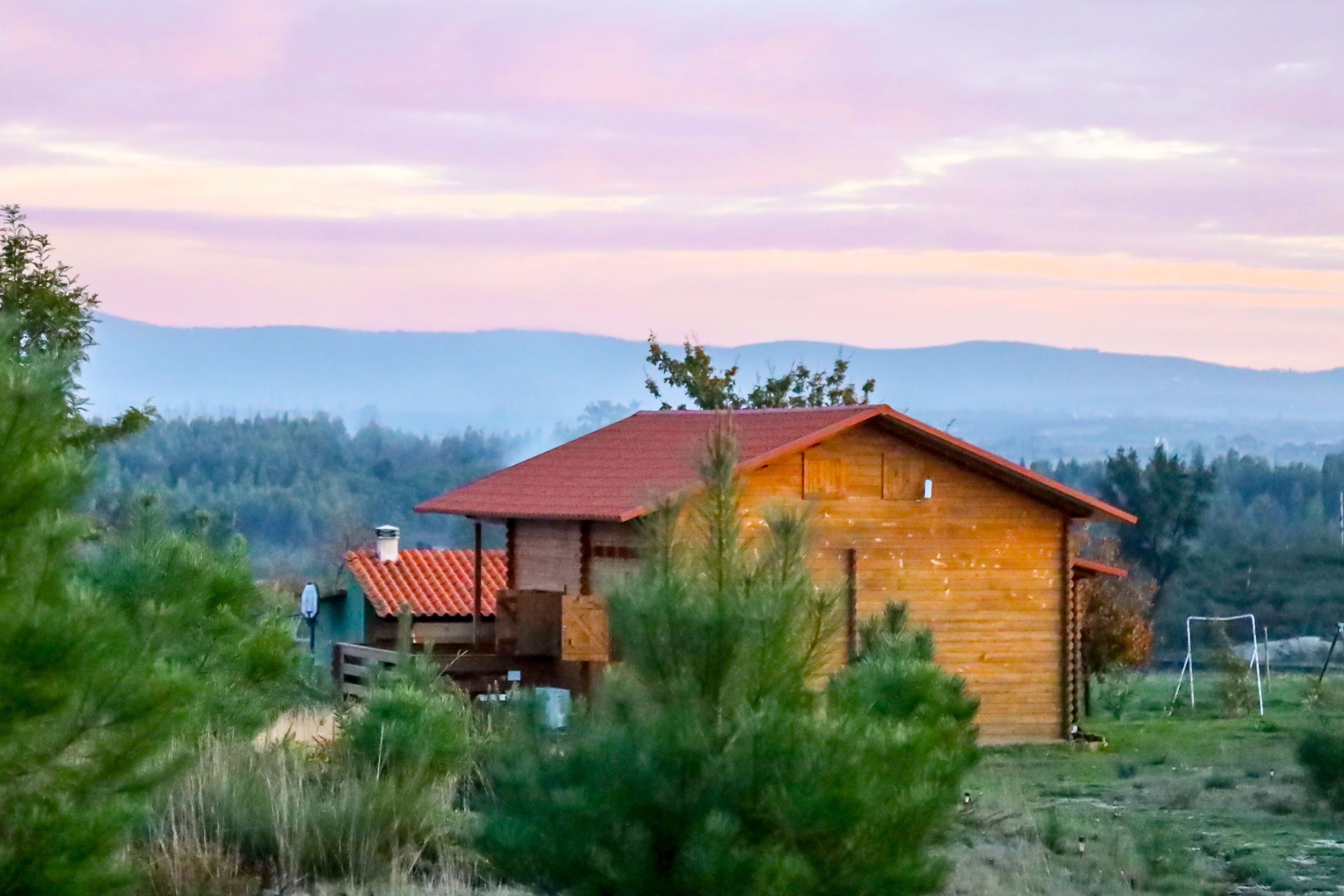 A wooden cabin with a red roof, surrounded by greenery and mountains in the background.