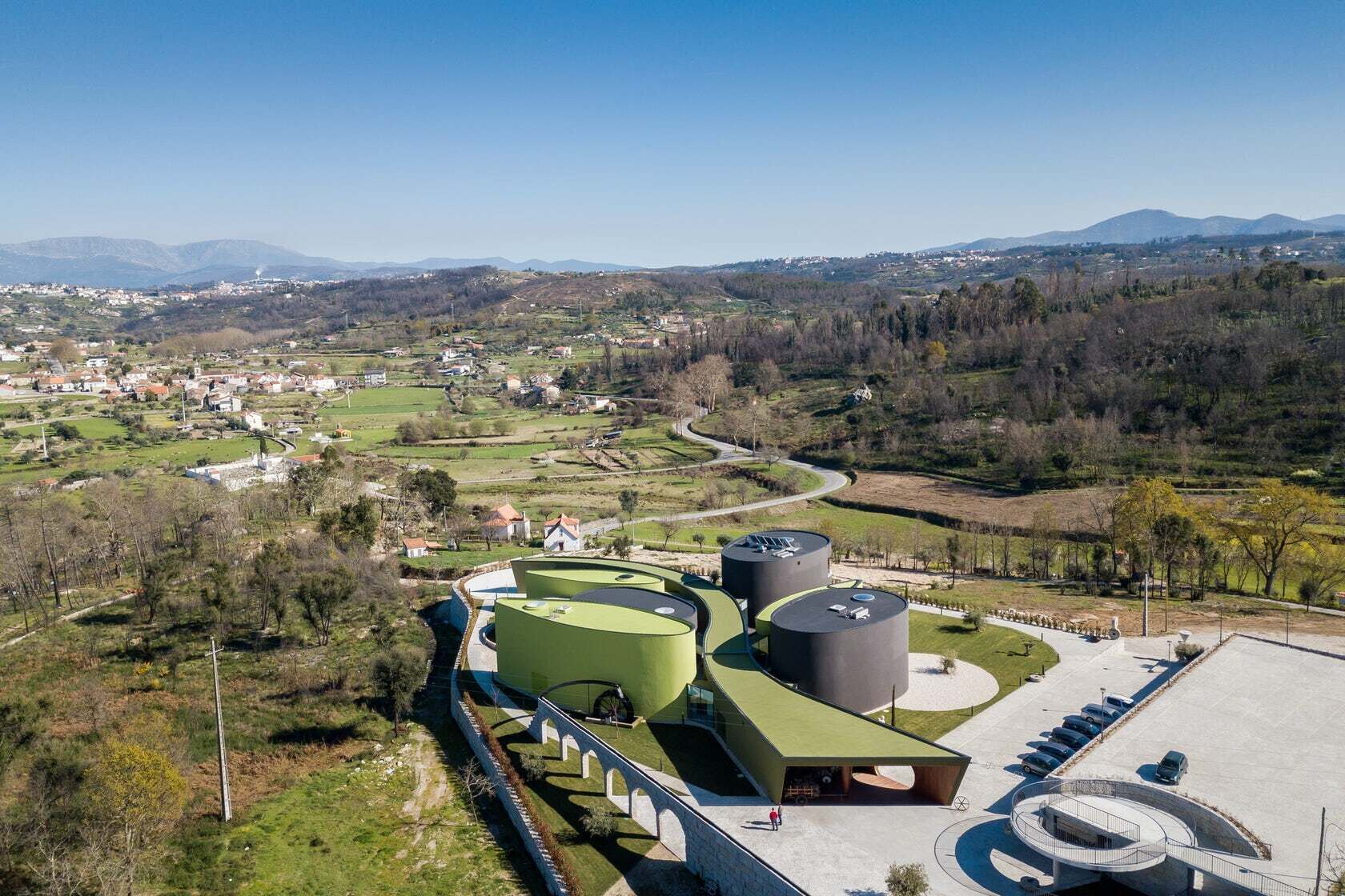 Un bâtiment moderne doté de toits végétalisés et de structures cylindriques, entouré d’un paysage rural avec des maisons et des montagnes en arrière-plan.