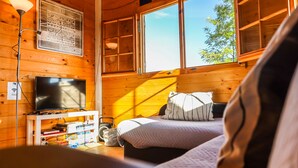 Una habitación acogedora tipo cabaña de madera con cama, televisor sobre un mueble bajo, estantería y ventana con vistas a los árboles.