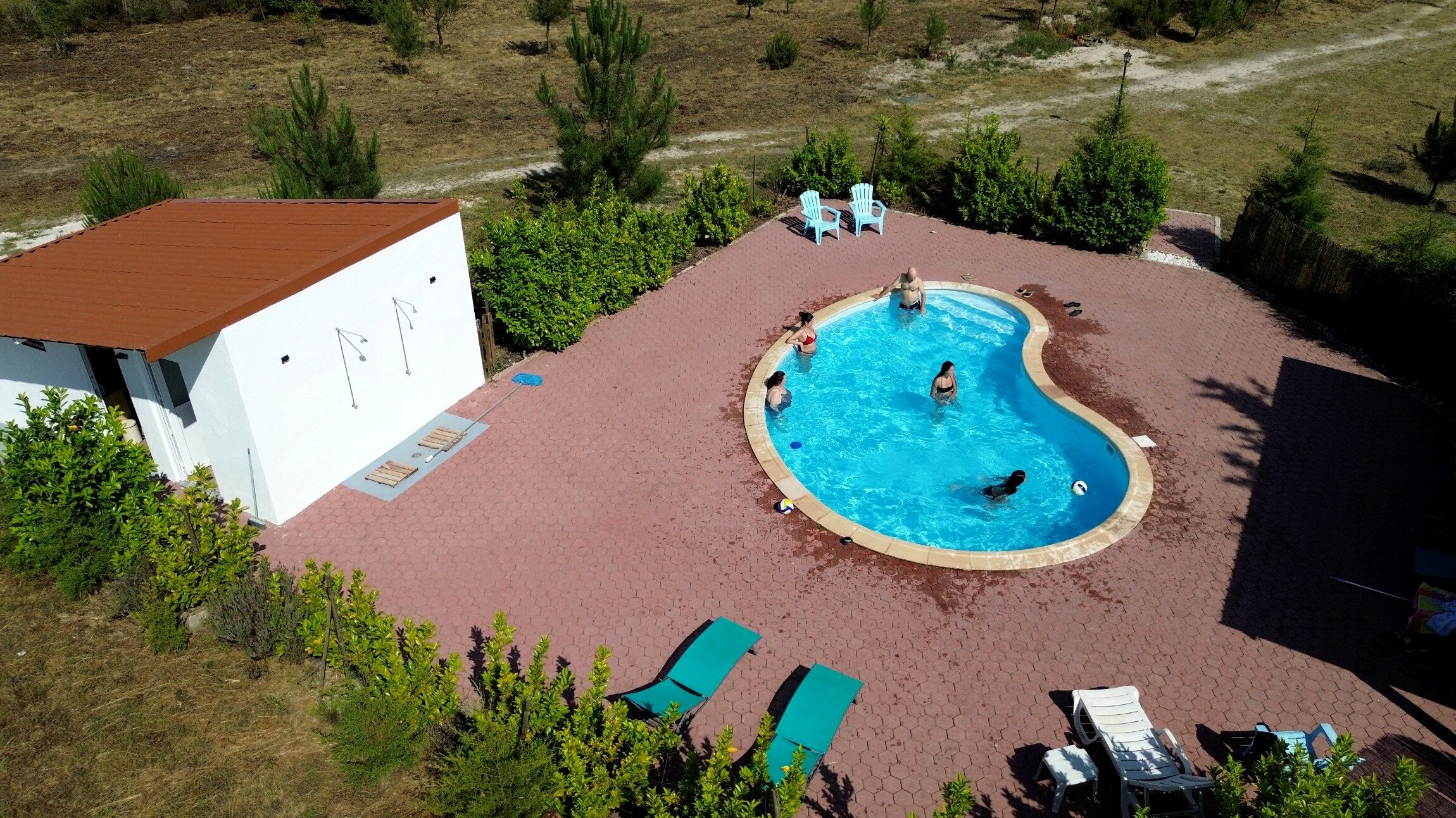 Une piscine extérieure à bords incurvés, entourée d’une zone dallée avec des chaises longues et un petit bâtiment doté d’un toit rouge.