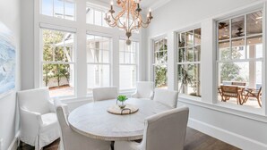 A dining area with a round table, white chairs, and a chandelier. Large windows provide views of the outdoors. - Luxury Home w/ Private Heated Pool in Seagrove! (Santa Rosa Beach)