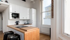 A modern kitchen with white cabinets, a wooden island, black stools, and a large window with a view of buildings. - Immaculate 1-Bed Apartment on Hove Seafront (Hove)