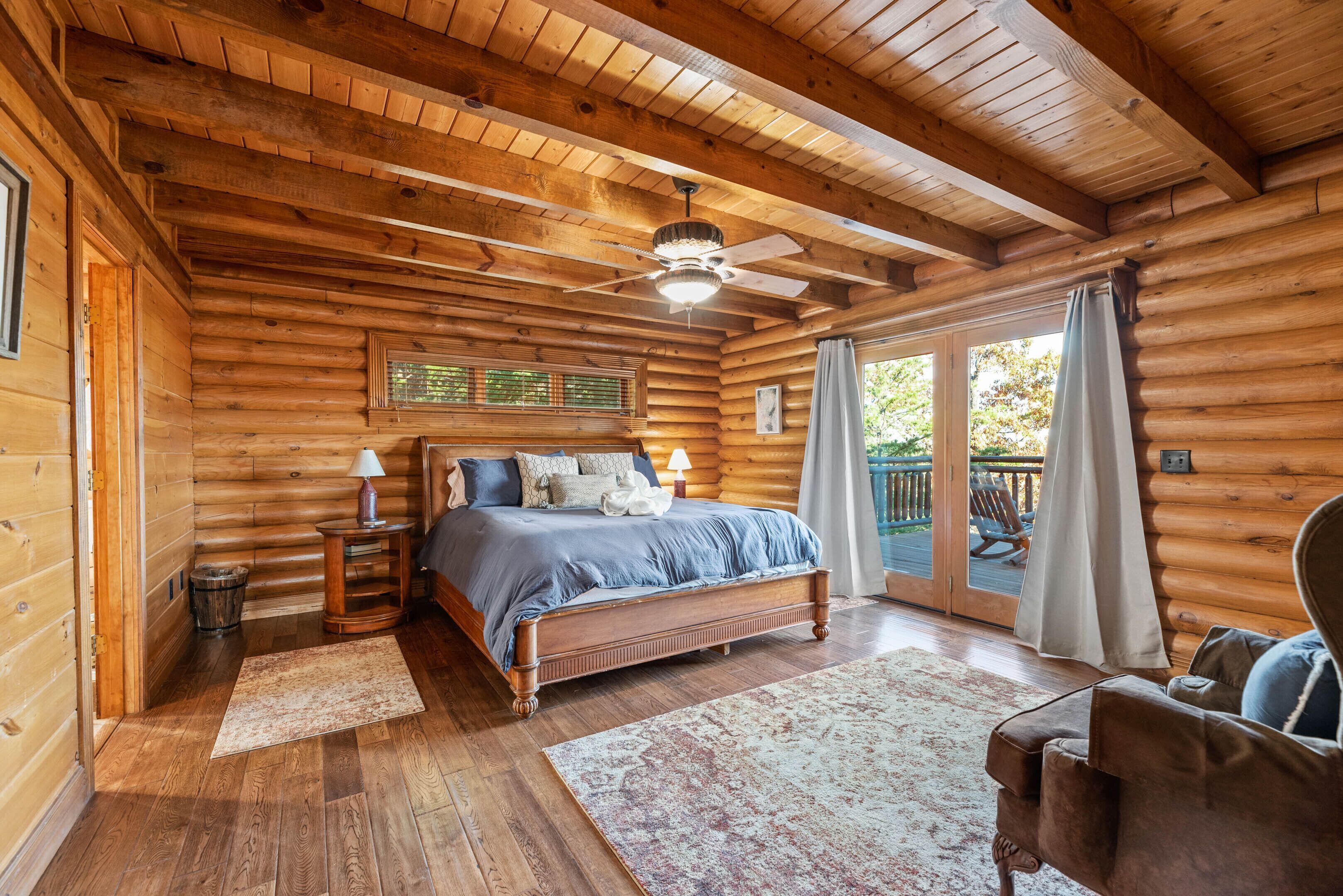 A log cabin bedroom with a wooden ceiling, a bed with blue bedding, a nightstand, a chair, and a view of the outdoors through a sliding glass door.