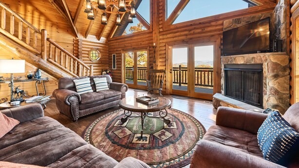 A spacious log cabin interior with a high ceiling, a fireplace, a large TV, and a cozy seating area with leather sofas and a round coffee table.