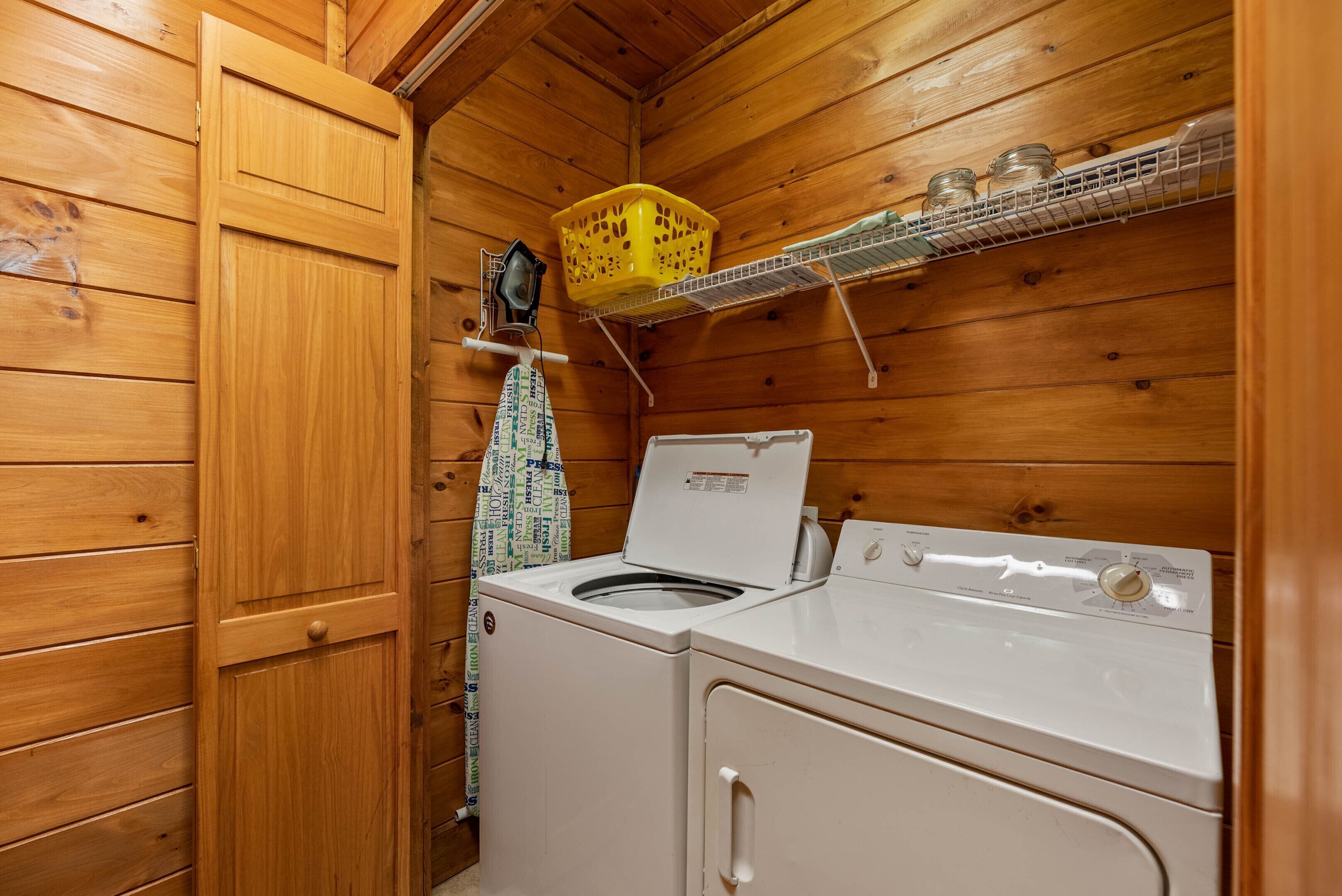 A compact laundry room with wooden walls, a washing machine, a dryer, a yellow basket, and a shelf with jars.