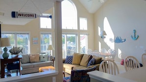 A spacious living area with a sofa, chairs, and a dining table. A 'WELCOME' sign hangs from the ceiling, and a 'LIVE LOVE LAKE' sign is visible on a window. - Modern Condo w/ Harbor views and Community Pool (Manistee)