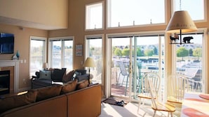 A spacious living room with a fireplace, a sofa, and a dining area with chairs. Large windows provide a view of the outdoors. - Modern Condo w/ Harbor views and Community Pool (Manistee)