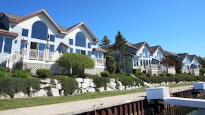 A row of white houses with balconies, a stone retaining wall, and a clear blue sky. - Modern Condo w/ Harbor views and Community Pool (Manistee)