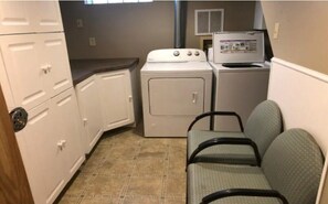 Laundry room with a front-loading washer and dryer, two chairs for waiting, and a small window for natural light.