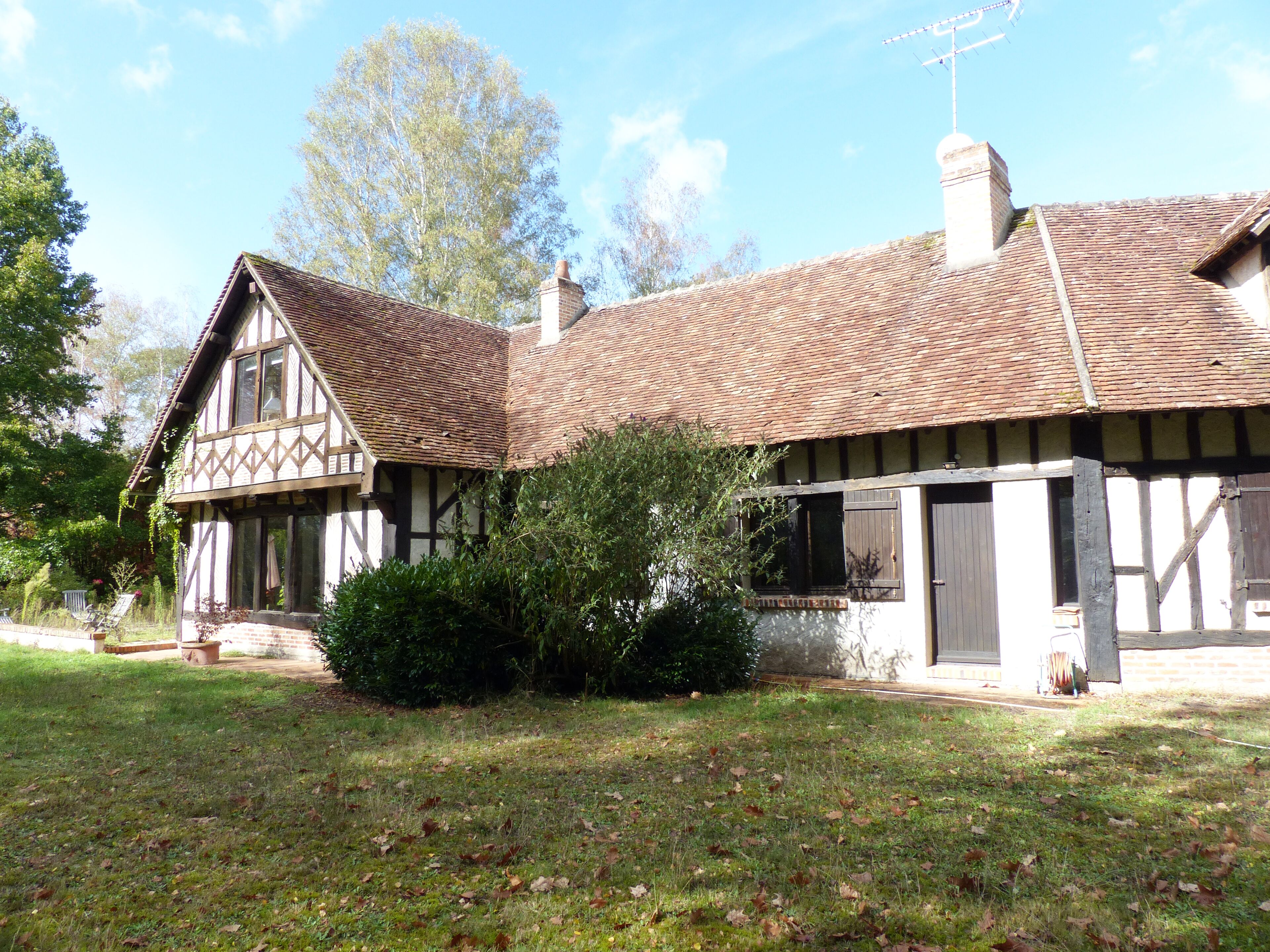 Une maison traditionnelle à colombages, dotée d’un toit en pente, d’une cheminée et d’un petit jardin agrémenté de pelouse et d’arbustes.