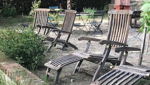 Ein Garten mit einer gepflasterten Terrasse, Holzstühlen und einem Tisch, umgeben von Grünpflanzen und einer Ziegelmauer.