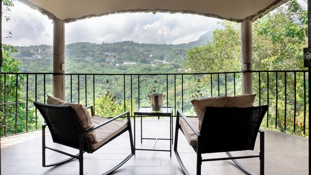 A balcony with two rocking chairs and a small table, overlooking a lush green landscape with hills and a town in the distance. - Hotel Paraiso Encantado (Xilitla)