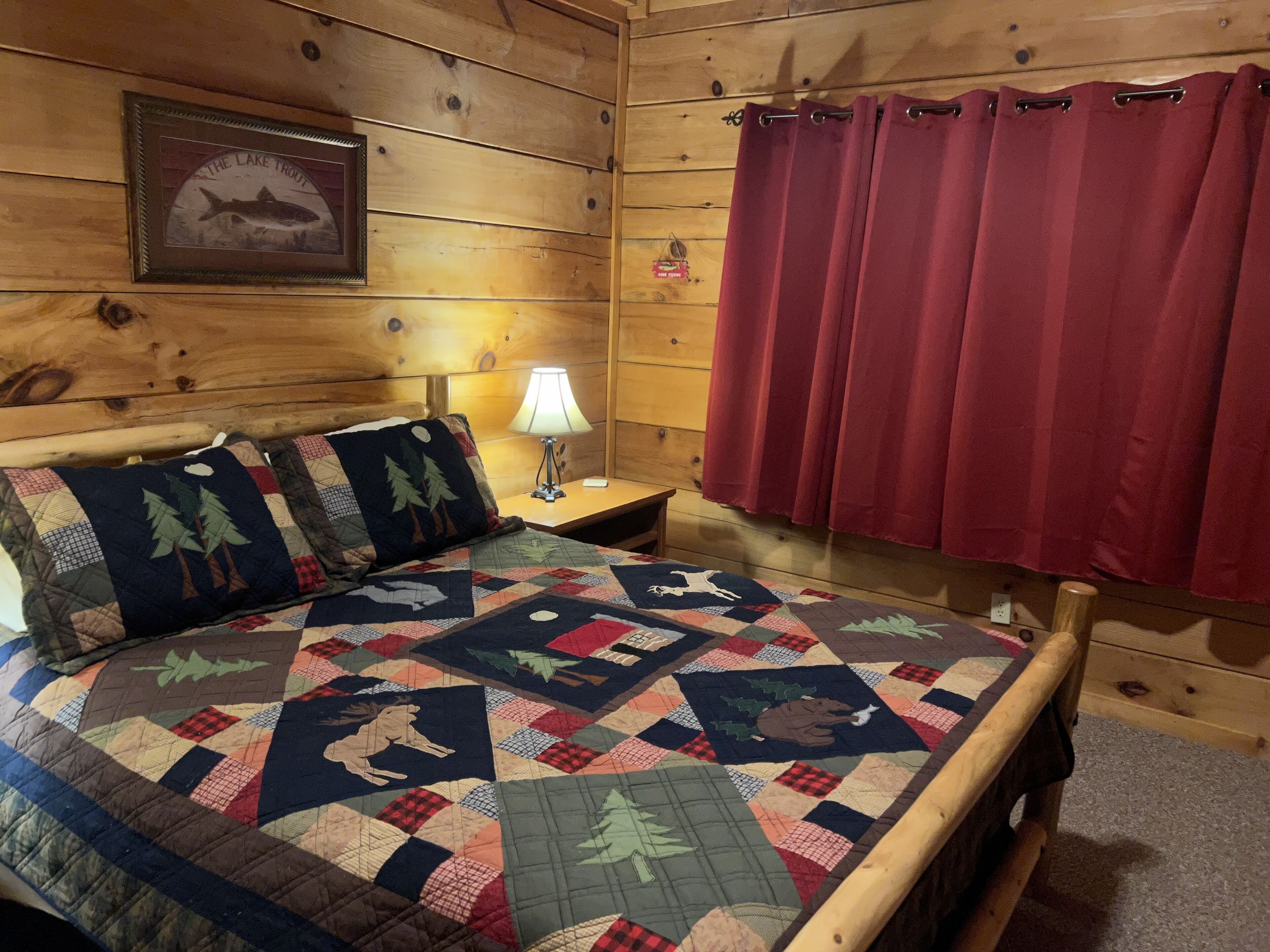 A wooden cabin room with a bed, a nightstand, a lamp, a red curtain, and a framed picture of a fish.
