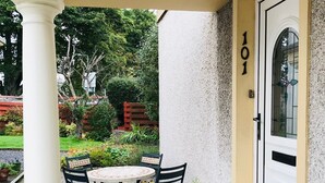 A covered patio with a round table and chairs, a white door with the number 101, and a garden with a red bench and potted plants. - Apartment 101 Kingsmills Road (Inverness)