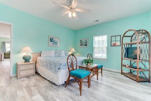 A bedroom with a bed, bedside tables, a desk with chairs, a TV on a wooden stand, and a ceiling fan.