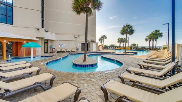 A pool area with lounge chairs, a palm tree, and a building with a tiled roof. - Sand Dunes Resort by Escape (Myrtle Beach)