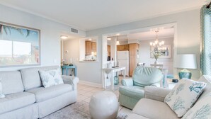 A spacious living room with a light-colored sofa, a round ottoman, and a teal armchair. A kitchen with wooden cabinets and a dining area with a white table and chairs are visible in the background. - Myrtle Beach Resort  (Myrtle Beach)