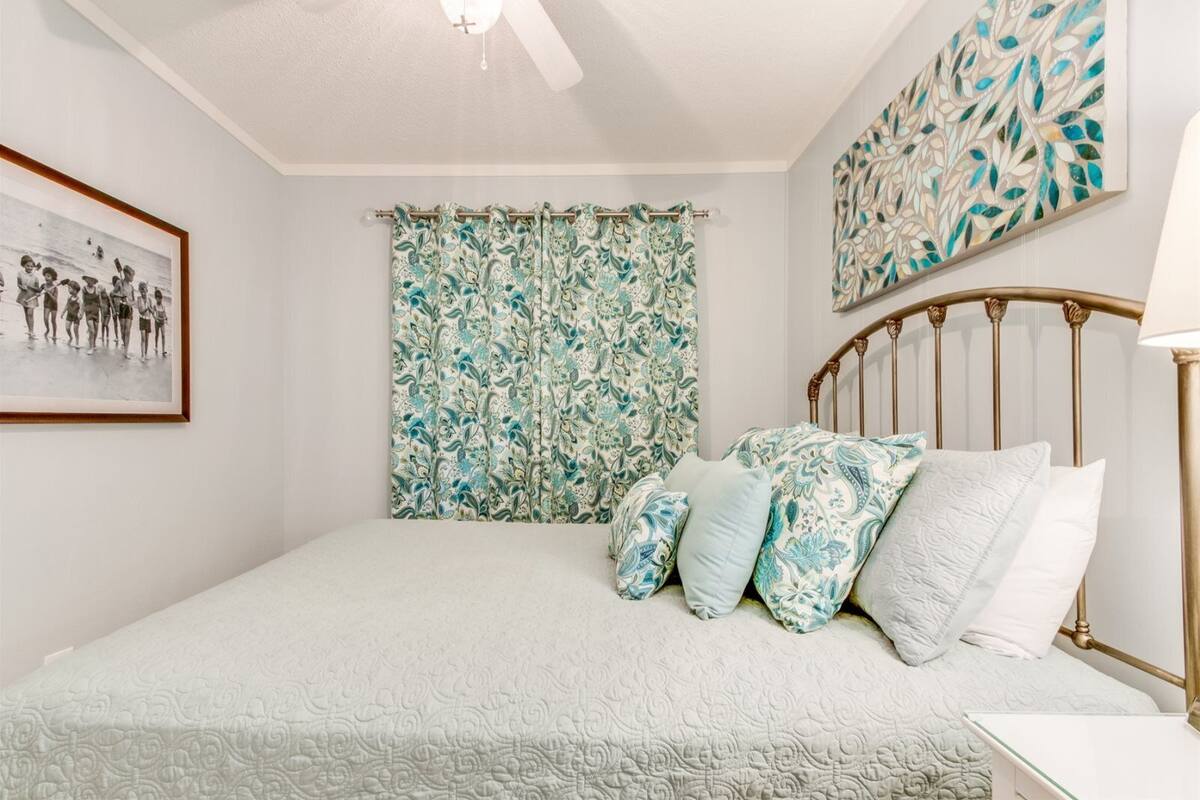 A bedroom with a bed, a ceiling fan, a framed picture, a floral curtain, and a wall art piece.