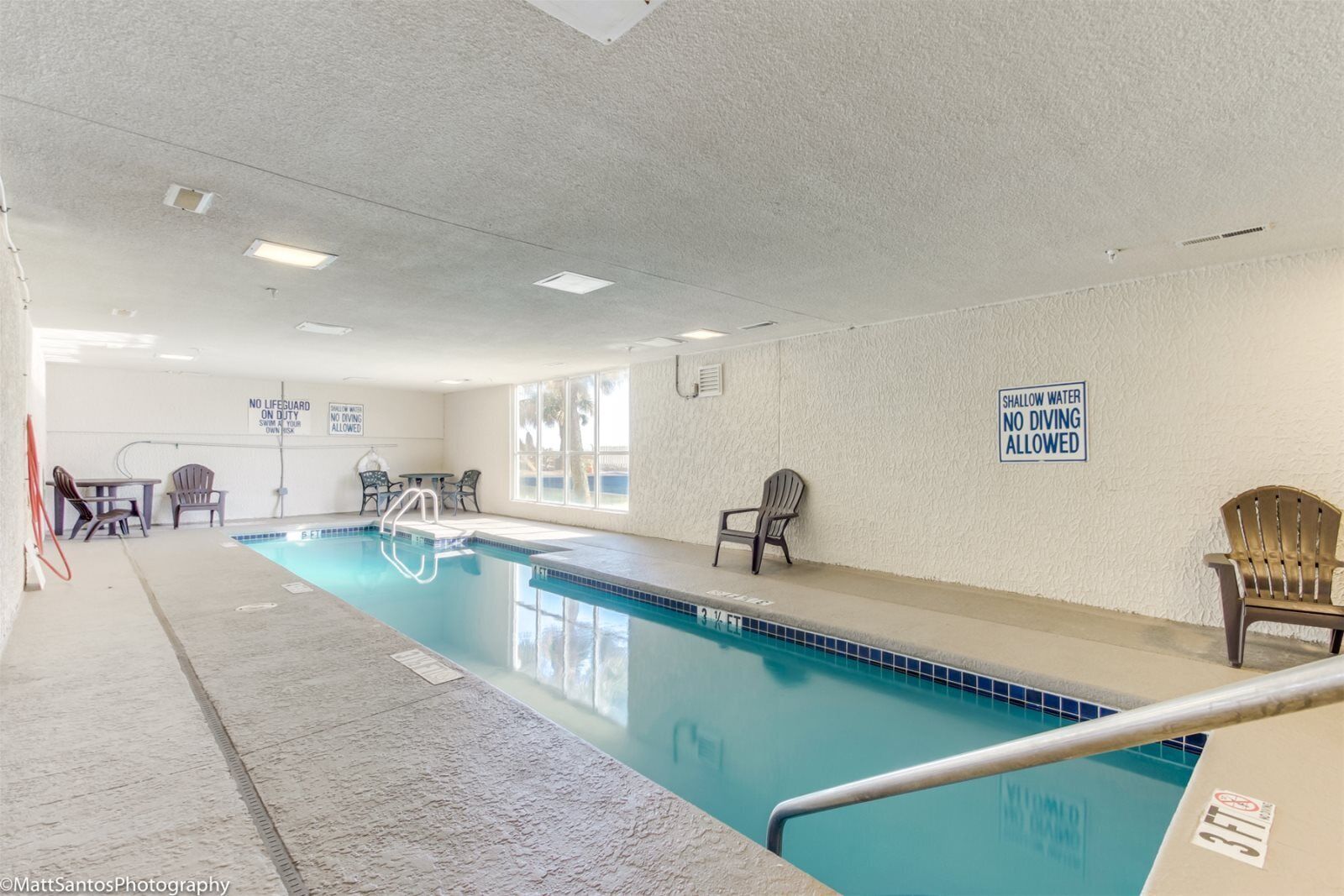 An indoor swimming pool with a shallow water sign, lounge chairs, and a lifeguard duty sign.