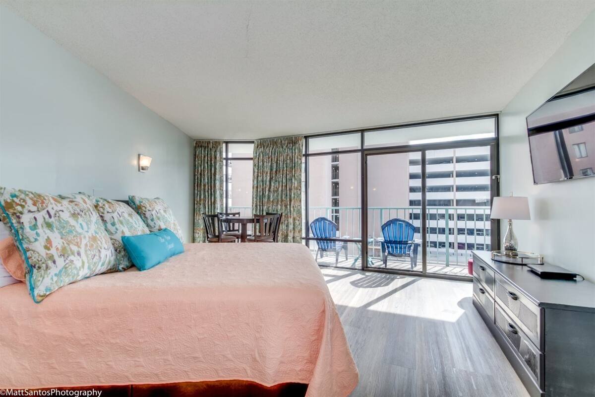 A hotel room with a large bed, a desk, and a balcony with chairs.