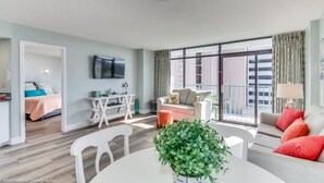 Eine moderne Wohnung mit Wohnzimmer, Essbereich und Schlafzimmer. Sie verfügt über ein großes Fenster, einen Flachbildfernseher und einen Balkon.