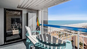 A balcony with a view of the beach, a poolside chair, and a door leading to a room. - Forest Dunes 1501 Escape! (Myrtle Beach)