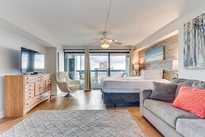 A modern hotel room with a bed, a sofa, a TV, a wooden dresser, and a balcony with a city view. - Forest Dunes 1501 Escape! (Myrtle Beach)