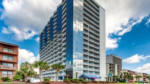 A modern high-rise hotel with multiple balconies, a glass facade, and a landscaped courtyard with palm trees. - Forest Dunes 1501 Escape! (Myrtle Beach)