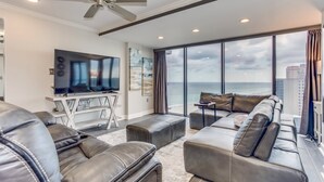 A modern living room with a large flat-screen TV, leather sofas, a ceiling fan, and a view of the ocean. - Forest Dunes 1501 Escape! (Myrtle Beach)