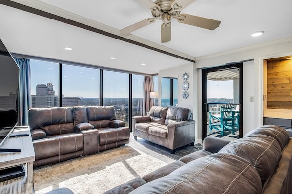 A spacious living room with leather sofas, a ceiling fan, and large windows offering a city view.