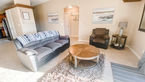 Een woonkamer met een grijze bank, een ronde salontafel, een bruine fauteuil en een bijzettafeltje met een lamp.