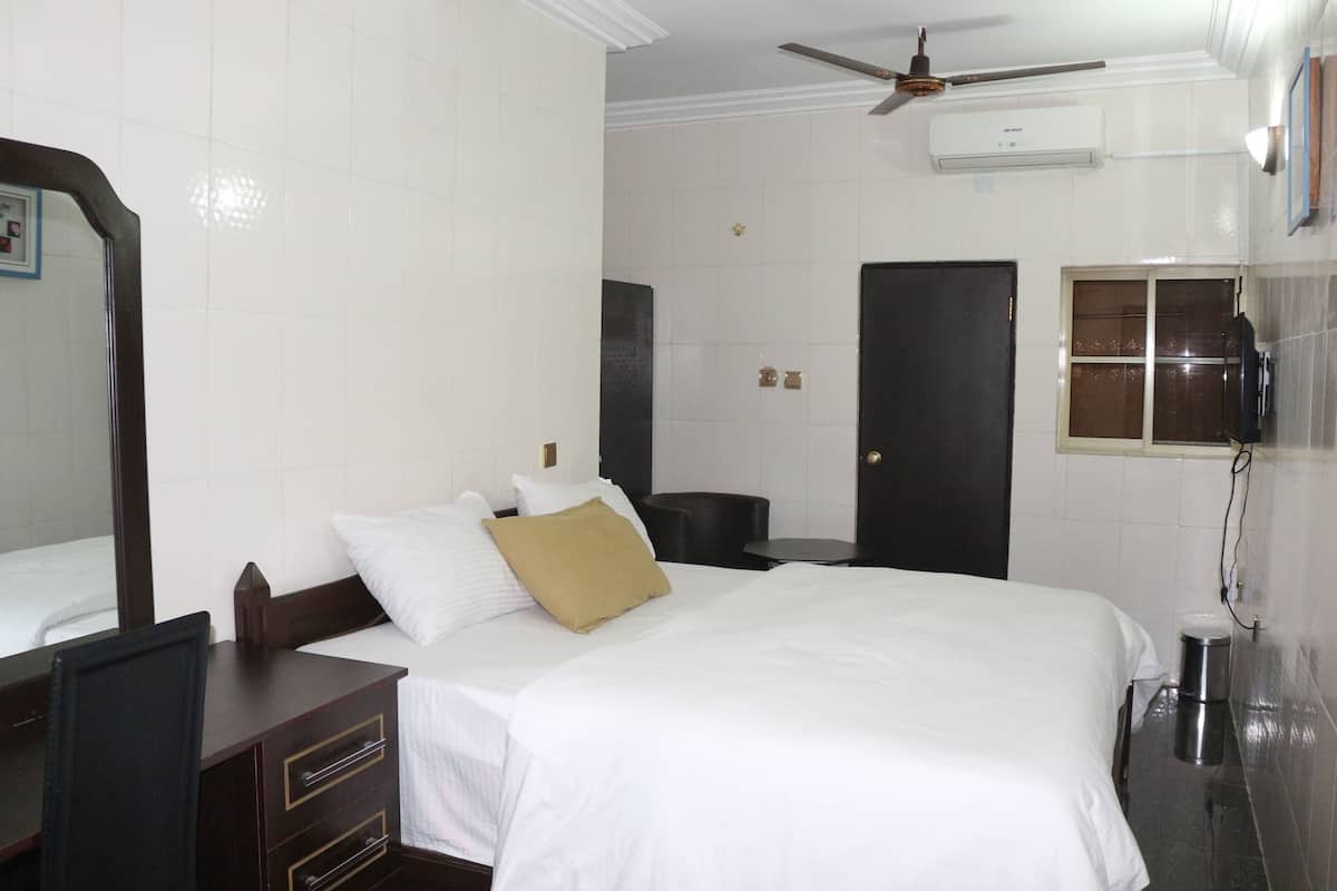 A hotel room with a bed, a desk, a chair, a wardrobe, a mirror, a ceiling fan, an air conditioning unit, a door, and a window.