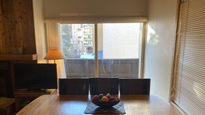 Ein Essbereich mit einem Holztisch, vier Stühlen und einer Obstschale. Ein Fernseher befindet sich links, und ein Fenster mit Jalousien rechts.