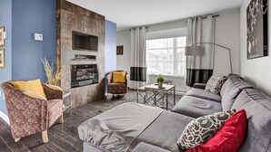 A modern living room with a fireplace, a sofa set, a patterned armchair, and a dining area with a table and chairs.