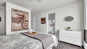 A modern bedroom with a large bed, a dresser, a mirror, and artwork on the walls.