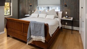 A bedroom with a wooden bed, a nightstand, a window with blinds, and a wall with vertical wooden slats.